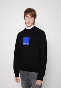 Felpa nera con polsini e orlo a costine, caratterizzata da un logo quadrato blu centrato sul petto. Il modello indossa jeans grigi.