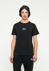 Crna pamučna majica s kratkim rukavima, okruglim izrezom, koja sadrži mali ispisani logotip na prsima. Ukupna glatka tekstura.