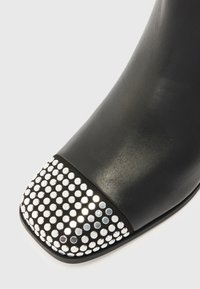 Must või nahast saapad, millel on ruudukujuline varvas ja kaunistatud läikivate hõbedaste ümarate nupudega esiserva ääres.