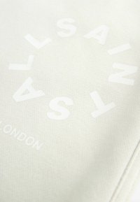 Plátno v jemné krémové barvě s výrazným, vyraženým bílým textem "SAINT LONDON." Hladký povrch s jemnými detaily prošívání.
