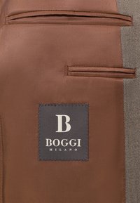 Tessuto marrone con una superficie liscia presenta due tasche angolate e un'etichetta con una "B" bianca su uno sfondo grigio scuro con la scritta "Boggi Milano".