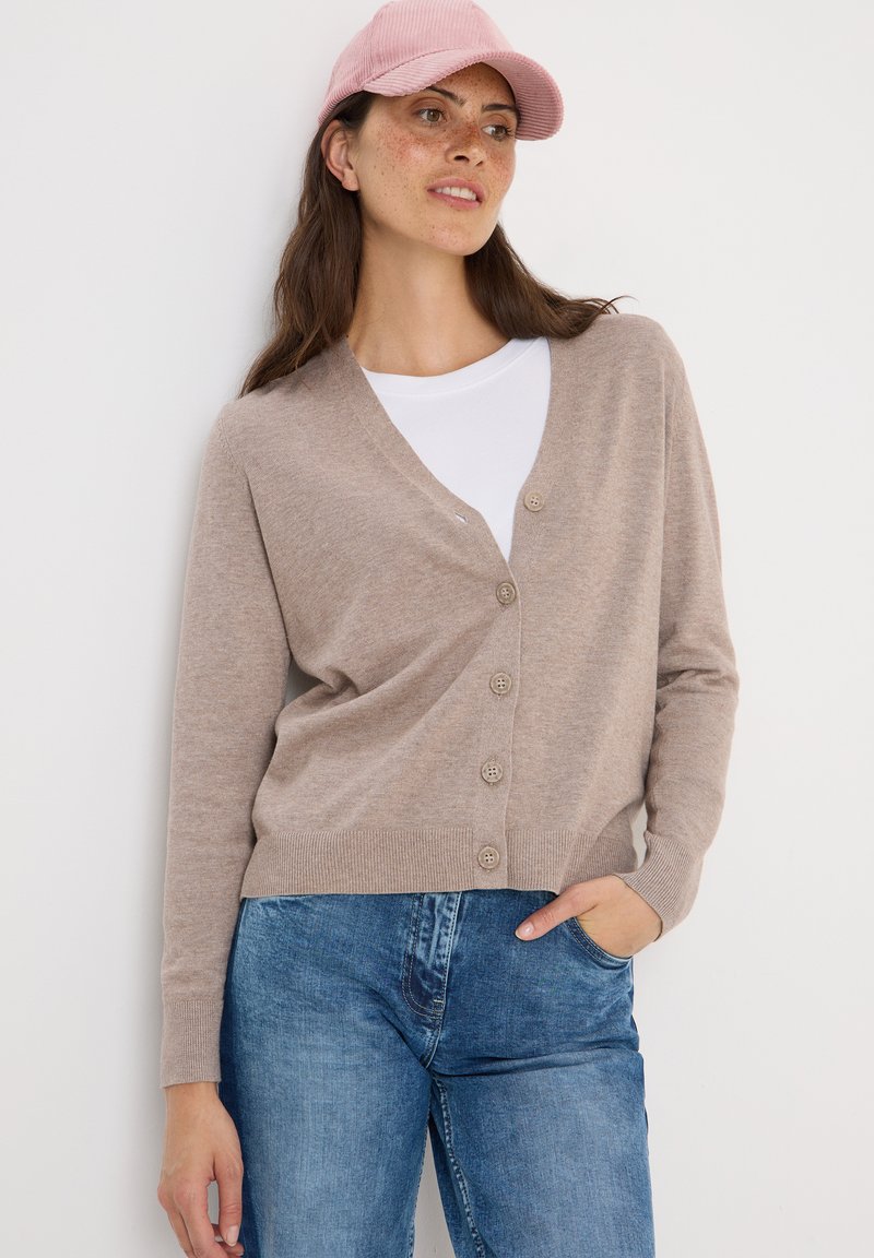 Beige cardigan med V-ringning, knappslå och ribbade muddarna, kombinerad med en vit T-shirt och denimjeans, bärs med en rosa keps.