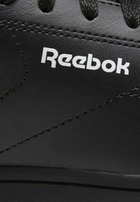 Černá sportovní obuv s hladkým koženým svškem, kulatými tkaničkami a bílým logem Reebok. Prošívání dodává designu texturu.