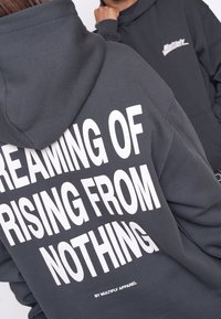 Felpa grigia scura con tasca frontale, caratterizzata da grande testo bianco: "SOGNANDO DI RIALZARSI DAL NULLA" e "DI MULTIPLY APPAREL."