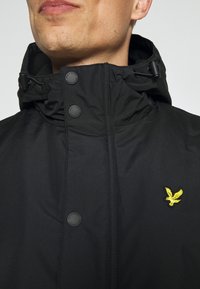 Czarna kurtka z wysokim kołnierzem, posiadająca czarne guziki oraz małe żółte logo orła na piersi. Gładki, wodoodporny materiał.