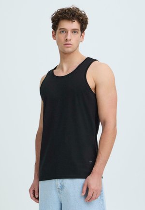 Ung mand med krøllet hår iført sort ærmeløs tanktop og lyseblå jeans, stående mod en ensfarvet lys baggrund.