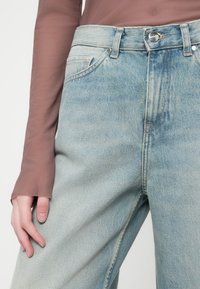 Svetlo modri denim hlače z visokim pasom, ki imajo pet žepov in subtilno drgnjenje. Dopolnjene z prosojno rjavo dolgimi rokavi.