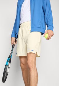 Shorts atletici de culoare bej deschis, cu talie elastică, fante laterale și un mic logo albastru. Persoana ține o rachetă de tenis și o minge de tenis verde.