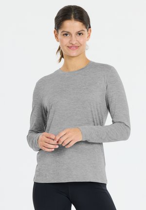 Kvinde med mørkt hår i en hestehale iført en ensfarvet grå langærmet trøje og sorte bukser, stående med hænderne let foldet.