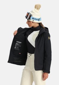 Schwarze Skijacke mit einem Reißverschluss vorne, der ein Logo sichtbar macht. Ein Paar weiße Skihosen und eine cremefarbene Mütze mit einem Bommel oben.