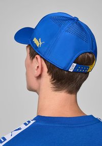 Niebieska czapka z zakrzywionym daszkiem, wykonana z gładkiego materiału, z żółtym logo Puma i perforowanymi detalami na górze. Regulowany pasek z tyłu.
