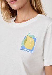 Biała bawełniana koszulka z grafika cytryny z zielonymi liśćmi i niebieskim tłem, z napisem "Zest d'Amour" pod wzorem.