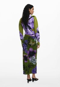 Gebreide groene trui met grote paarse bloemenprint; gecombineerd met een vloerlange rok met vergelijkbare bloemenpatronen in groen en paars.