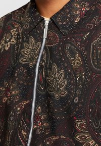 Close-up van donker paisley-gestoffeerd met tinten rood, beige en groen, met een zilveren rits en een geklede halslijn.