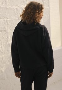 Schwarzer Kapuzenpullover mit lockerer Passform, ausgestattet mit einer sanften Textur und gerippten Bündchen. Der Stoff wirkt dick und warm, ideal für die Freizeit.