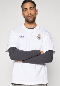 T-shirt branca de algodão com mangas curtas, apresentando um logótipo roxo da Adidas e o emblema do Real Madrid. Vestida por cima de uma camisa cinzenta de manga comprida.