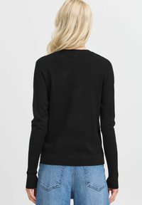 Maglione nero a maglia con maniche lunghe, polsini e orlo a costine. Indossato sopra jeans blu a vita alta, presenta un design semplice e classico.