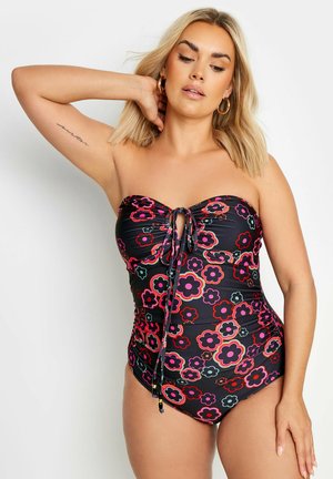Blondynka w czarnym, bezramkowym kostiumie kąpielowym jednoczęściowym z kolorowym kwiatowym wzorem i wiązaniem z przodu, pozująca z uniesionym ramieniem.