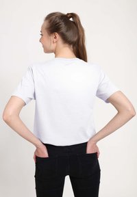 Biała koszulka z krótkim rękawem i okrągłym dekoltem, o gładkim wzorze. Model nosi czarne dżinsy, ręce w kieszeniach, pokazany od tyłu.