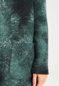 Cappotto misto lana in verde scuro con una finitura testurizzata e marmorizzata. Presenta una tasca laterale e un polsino della manica aderente.