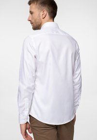 Biała koszula z długim rękawem i klasycznym kołnierzykiem, gładka w dotyku, z zaokrąglonym dołem i mankietami na guziki. Noszona z beżowymi spodniami, widok z tyłu.