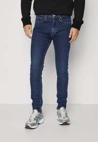 Jeans in denim blu skinny, con un classico design a cinque tasche, hardware in rame e una texture liscia. Abbinati a delle scarpe da ginnastica argentate.