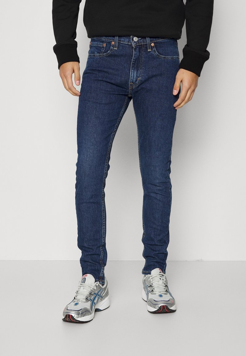 Dunne blauwe denim jeans met een klassiek vijf-pocket ontwerp, koperen hardware en een gladde textuur. Gepaard met zilveren sportschoenen.