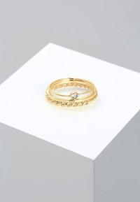 Set di anelli in tono oro caratterizzati da un design intrecciato e una piccola pietra incastonata, esposti su un piedistallo bianco con uno sfondo pulito e minimale.