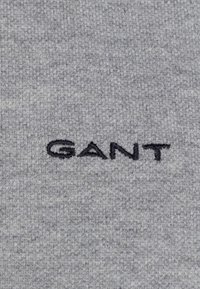 Černé logo "GANT" vyšívané na světle šedém pleteném pozadí.
