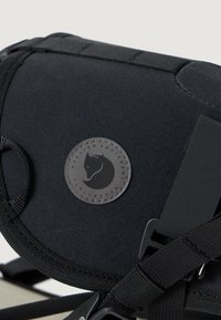 Svart stoff skulderveske med en sirkulær logo-patch, justerbare stropper og webbing for sikker festing. Glatt tekstur og kompakt form.