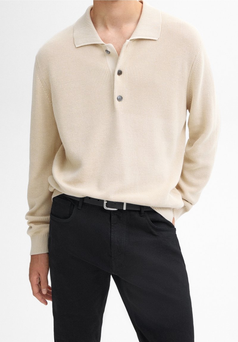 Homme portant un polo en maille beige à manches longues avec trois boutons et un pantalon noir avec une ceinture noire sur fond blanc.