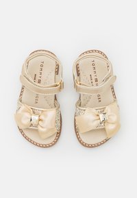 Sandali dorati con una tomaia testurizzata, caratterizzati da un grande fiocco e dettagli glitterati. Cinturino regolabile con hardware metallico, suola piatta e bordi cuciti.