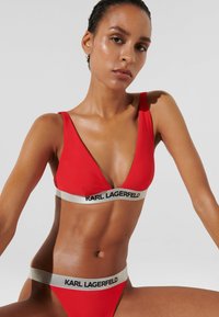 Crveni bikini set s dubokim V-izrezom na gornjem dijelu i odgovarajućim niskim donjim dijelom. Svaki komad ima kontrastni crni i srebrni pojas s logom "KARL LAGERFELD".