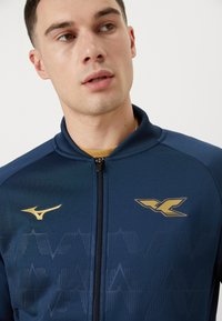 Veste de sport bleu marine avec fermeture éclair, dotée de accents dorés et de motifs géométriques texturés sur le tissu. Conception à col haut.