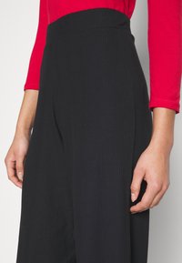 Černé široké kalhoty s žebrovaným vzorem a elastickým pasem. Částečně viditelná je červená dlouhá košile s přiléhavým střihem.