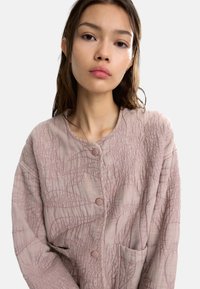 Teksturowana, jasnoróżowa kurtka z okrągłym dekoltem, zapięciem na guziki i dwiema przednimi kieszeniami. Charakteryzuje się wyraźnymi przeszytymi wzorami.
