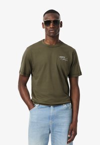 Camisa de algodón color verde oliva con cuello redondo y mangas cortas, que presenta un pequeño logo bordado en el lado izquierdo del pecho. Llevada con jeans de mezclilla azul claro.