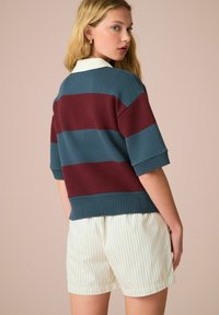 Sweter polo z dzianiny w poziome paski w odcieniach turkusu i burgunda, z białym kołnierzykiem. Połączony z kremowymi spodniami w paski.