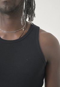 Schwarzes geripptes Tanktop mit Rundhalsausschnitt, das eine dezente Logo-Stickerei aufweist. Der Stoff scheint weich zu sein und hat ein tailliertes Design. Eine silberne Kettenhalskette ist sichtbar.