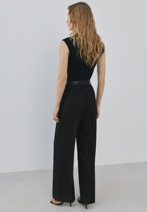 Frau mit langen, welligen Haaren steht mit dem Gesicht zur Wand, trägt ein ärmelloses schwarzes Oberteil, schwarze weitgeschnittene Hose, schwarzen Gürtel und schwarze Sandalen mit Absatz.