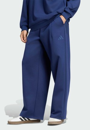 Persona que lleva pantalones sueltos azul oscuro de Adidas y sudadera a juego con zapatillas blancas con rayas negras.
