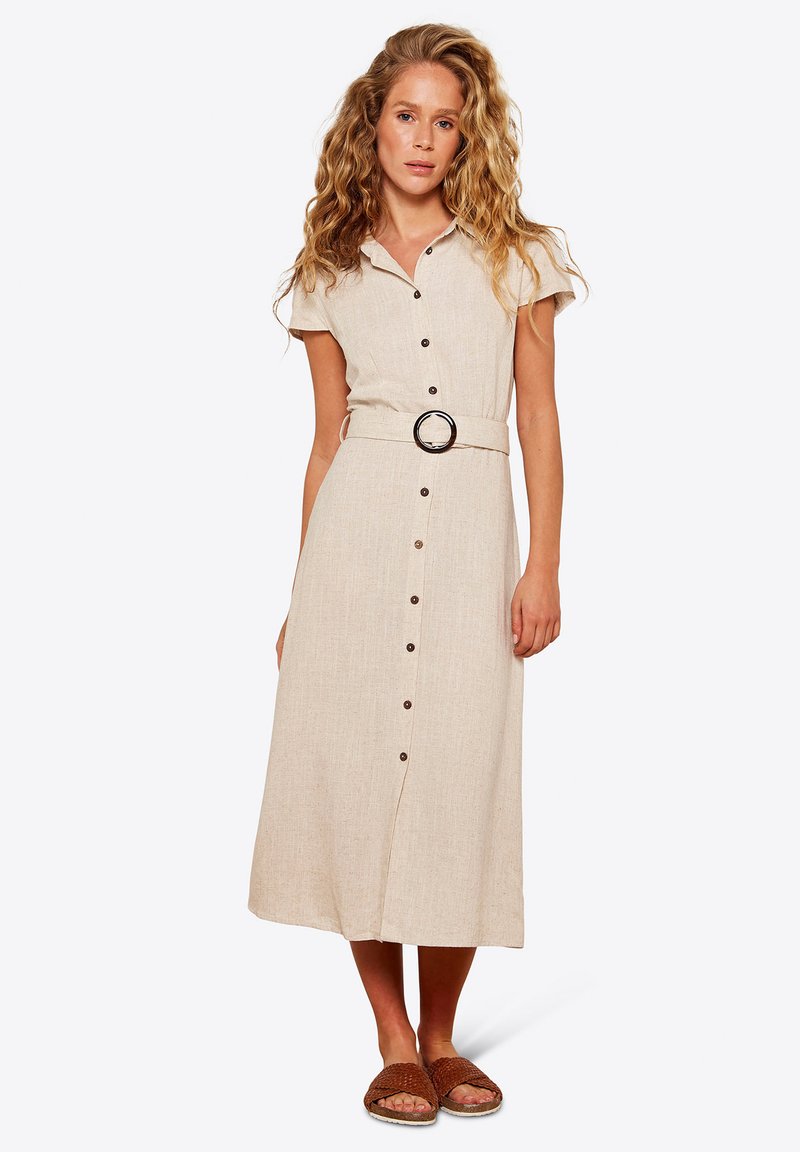 Beige Leinenkleid mit kurzen Ärmeln, Knopfleiste und kreisförmigem Gürtelakzent. Glatte Textur, knielanger Rock, getragen mit braunen Rutschsandalen.