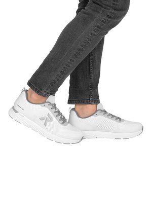 Person, die weiße und graue Sportschuhe mit grauen Schnürsenkeln und dunkelgraue Jeans trägt, steht mit einem leicht angehobenen Fuß.