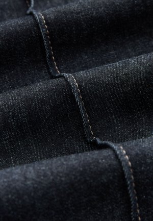 Tela de mezclilla negra con una textura suave y un brillo sutil. Presenta costuras en contraste de color marrón claro y patrones de tejido diagonal tenue.