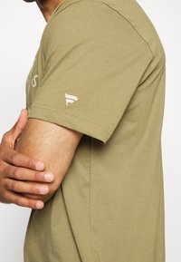 Bavlněné tričko olivově zelené barvy s krátkými rukávy. Na levém rukávu je malý logo a střih je volný. Hladká textura.