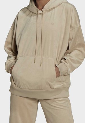 Personne portant un sweat à capuche en velours beige et un pantalon assorti, les mains dans la poche avant du sweat, montrée du cou jusqu'au milieu de la cuisse.