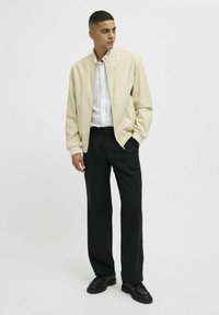 Veste bomber beige clair, fermeture éclair à l'avant, poignets à côtes, associée à une chemise blanche à boutons et un pantalon noir. Tissu lisse au design discret.