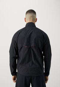 Veste légère noire avec des manches longues et un col haut. Comprend un détail découpé horizontal à l'arrière et un tissu texturé.