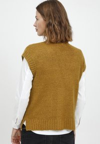 Senfgelbe, gestrickte, ärmellose Sweater mit strukturiertem Finish, geripptem Saum und Seitenschlitzen, über einem weißen Langarmshirt getragen.