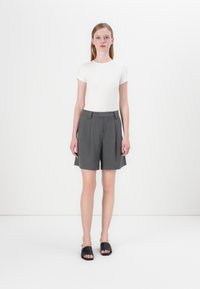 Grå plissert shorts med skreddersydd passform og lommer, sammen med en tettsittende hvit T-skjorte og svarte sandaler. Glatt stofftekstur.